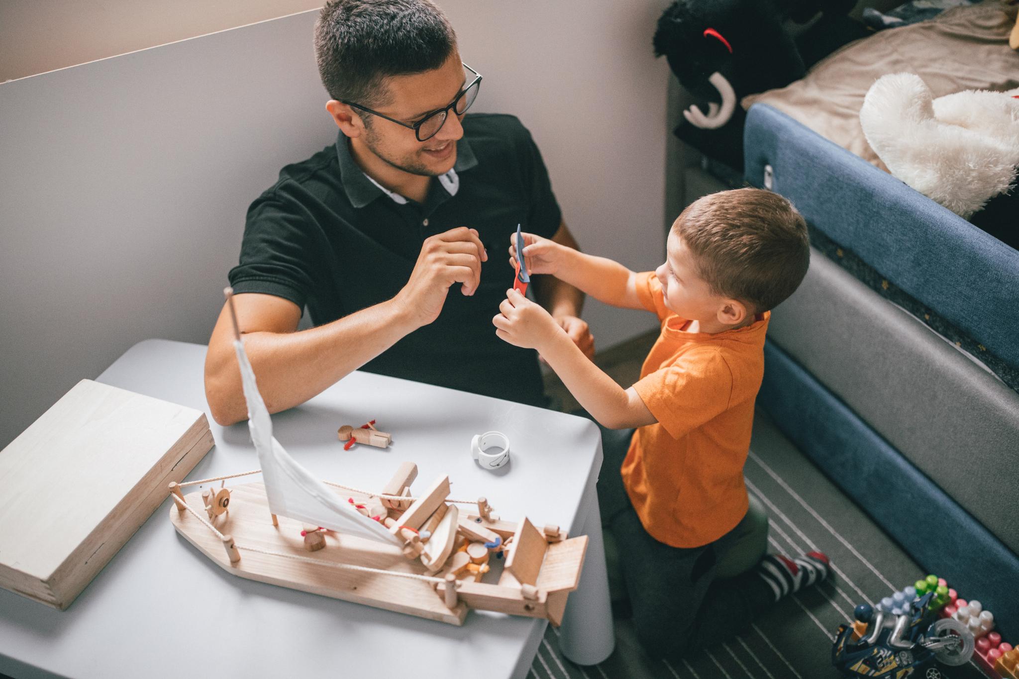 Ein Mann und ein Kind knien auf dem Boden zwischen einem Bett und einem Tisch. Der Mann hat dunkles, kurzes Haar, ein schwarzes Hemd und eine schwarze Brille, und das Kind hat helleres, kurzes Haar und ein orangefarbenes T-Shirt. Auf dem Tisch liegt ein Holzspielzeugschiff. Das Kind zeigt dem Mann einige kleine Zettel.