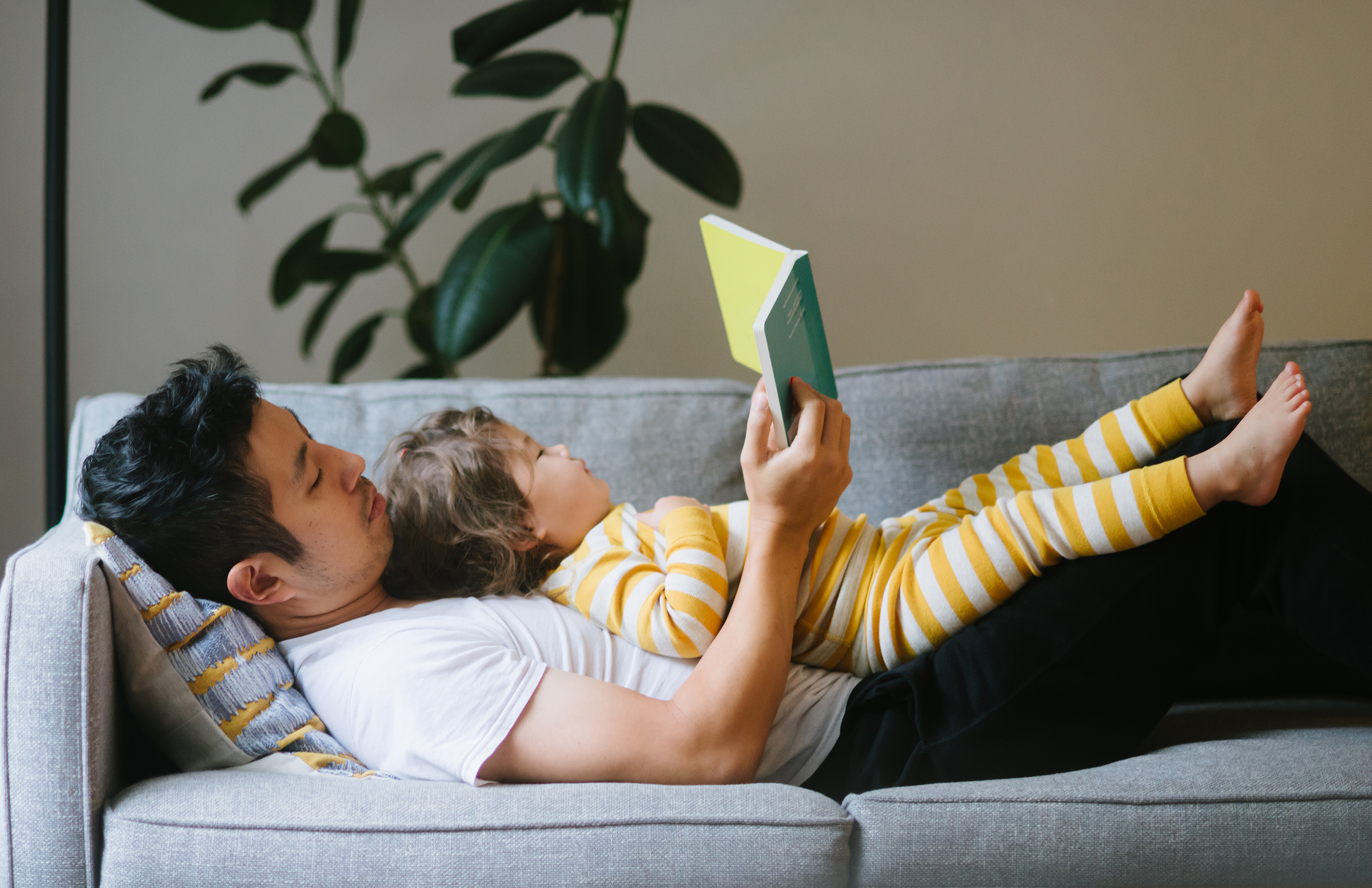 Ein Mann mit kurzen, glatten, schwarzen Haaren liegt auf einem grauen Sofa, auf ihm liegt ein Kleinkind. Das Kleinkind hat mittellanges braunes Haar und trägt einen gelb-weiß gestreiften Schlafanzug. Sie lesen gemeinsam ein Buch.